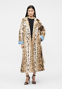 Femeie purtând o haină lungă cu imprimeu de leopard și manșete albastre peste o rochie neagră, asortată cu sandale negre cu toc înalt și cercei pendanți.