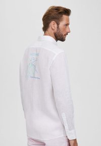 Camisa de lino blanca con un diseño de bordado en la parte posterior que presenta acentos coloridos y el texto "FAÇONNABLE". Mangas largas con puños abotonados.