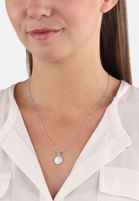 Collar de cadena de plata con un colgante redondo de superficie texturizada. El colgante se encuentra sobre una blusa de color claro.