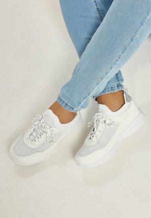 Person wearing light blue jeans and white mesh sneakers with laces, sitting with legs crossed against a light background.