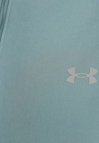 Hellblauer sportlicher Stoff mit glatter Textur und einem dezenten Under Armour-Logo in einem etwas dunkleren Farbton. Reißverschlussdetail oben sichtbar.