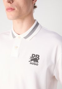Witte poloshirt met een tweeknoopsgat, grijs gestreepte kraag en een zwart logo met een stier en de tekst "PR HUGO." Glad textuur.