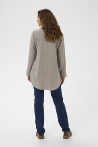 Kvinna med vågigt brunt hår står med ryggen mot kameran, klädd i en ljusgrå långärmad skjorta, mörkblå jeans och bruna skor mot en vit bakgrund.