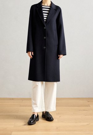 Personne portant un long manteau noir boutonné, un haut rayé noir et blanc, un pantalon blanc à jambes larges et des mocassins noirs debout sur un sol en bois.