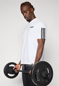 Witte katoenen sportshirt met zwart logo, voorzien van drie zwarte strepen op de linkermouw. Model houdt een halter met zwarte gewichten vast.