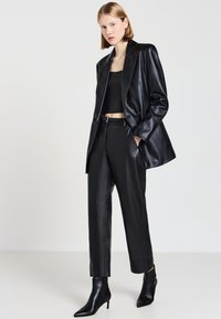 Schwarze Lederblazer über einem schwarzen Crop-Top, kombiniert mit schwarzen Lederhosen und spitzen Ankle Boots. Glatte Textur, maßgeschneiderte Passform.