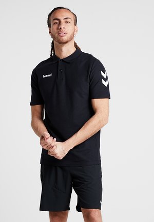 T-shirt polo noir avec un col, portant un logo blanc sur la poitrine gauche et des accents en chevron blancs sur les manches. Porté avec un short noir.