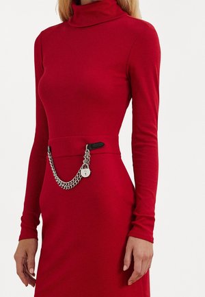 Nainen, joka pukeutunut istuvaan punaiseen poolomekkoon, jossa on pitkähihaiset hihat ja hopeinen ketjuvyö, jossa on lukon yksityiskohta.