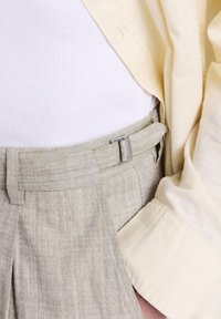 Pantaloni di lino beige con fibbia laterale, top bianco a coste e camicia a maniche lunghe gialla chiaro con la mano in tasca.