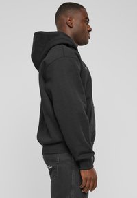 Sudadera negra con capucha y bolsillo tipo canguro, con puños y dobladillo acanalados, que presenta un pequeño logo blanco en el lado izquierdo del pecho. Textura de tela suave.