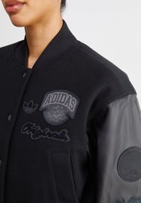 Zwarte varsityjack met een wollen lichaam, leren mouwen en geborduurde logo's. Voorzien van een geribde kraag en een sluiting met drukknoop aan de voorkant.