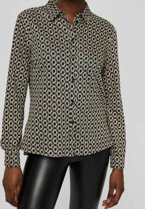 Persoon die een overhemd met lange mouwen draagt met een zwart-beige geometrisch patroon en een zwarte leren broek, staand tegen een effen achtergrond.