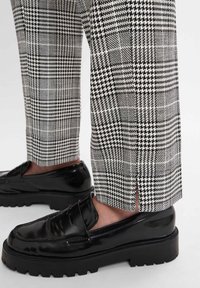 Schwarz-weiße karierte Hose mit geradem Schnitt, mit einem seitlichen Schlitz am Saum, kombiniert mit schwarzen glänzenden Slippern auf einer dicken Sohle.