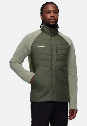 Homme portant une veste zippée Mammut olive avec un devant matelassé et des manches en polaire, se tenant avec une main dans la poche.