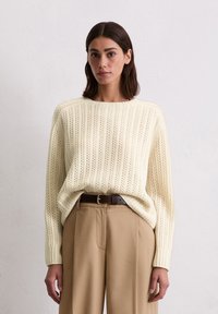 Cremiger Strickpullover mit vertikaler Rippung und offener Webstruktur, kombiniert mit hochschnittigen, beigen Hosen und einem braunen Gürtel.