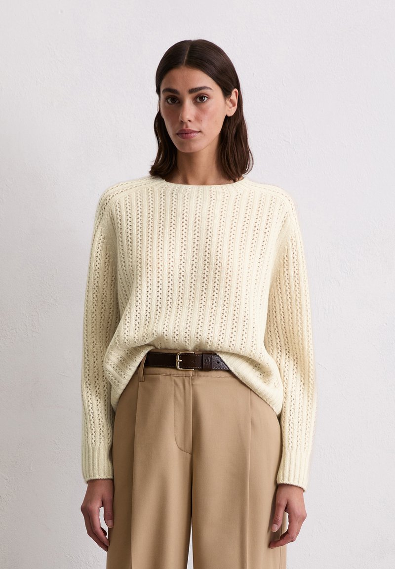 Cremiger Strickpullover mit vertikaler Rippung und offener Webstruktur, kombiniert mit hochschnittigen, beigen Hosen und einem braunen Gürtel.