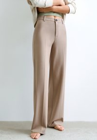 Beige leveled trousers made from smooth fabric, featuring a button closure and a tailored fit. They fall to the floor with a clean hem.