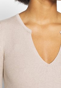 Béžový žebrovaný pletený top s hlubokým výstřihem do V a neupravenými okraji u výstřihu. Materiál je jemný a měkký na omak.
