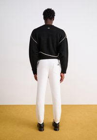 Sort cropped sweater med creme detaljer, parret med hvide tapered bukser og sorte chunky sko. Ensfarvet baggrund, gul gulv.