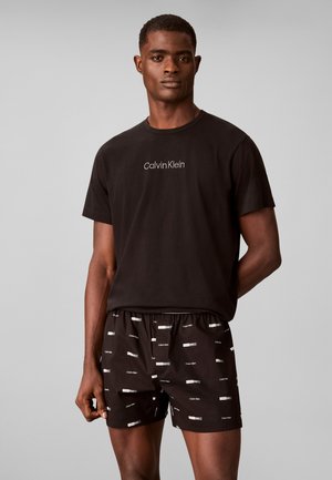 Мъж, облечен в черна тениска Calvin Klein и черни къси панталони с малки бели лога на Calvin Klein, стоящ на сив едноцветен фон.