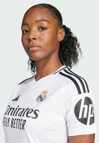 White soccer jersey with black accents, featuring "Emirates Fly Better" text, Real Madrid logo, and HP logo on the sleeve. Textured fabric design.