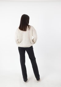 Jasno beżowy, oversize'owy sweter dzianinowy z opadającymi ramionami, zestawiony z dopasowanymi, czarnymi dżinsami, widziany z tyłu na tle gładkiej białej ściany.