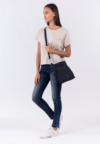 Borsa a tracolla nera con superficie texturizzata e decorazione con nappina. Il modello indossa una maglietta beige chiaro, jeans scuri con dettagli in zip blu e sneakers bianche.