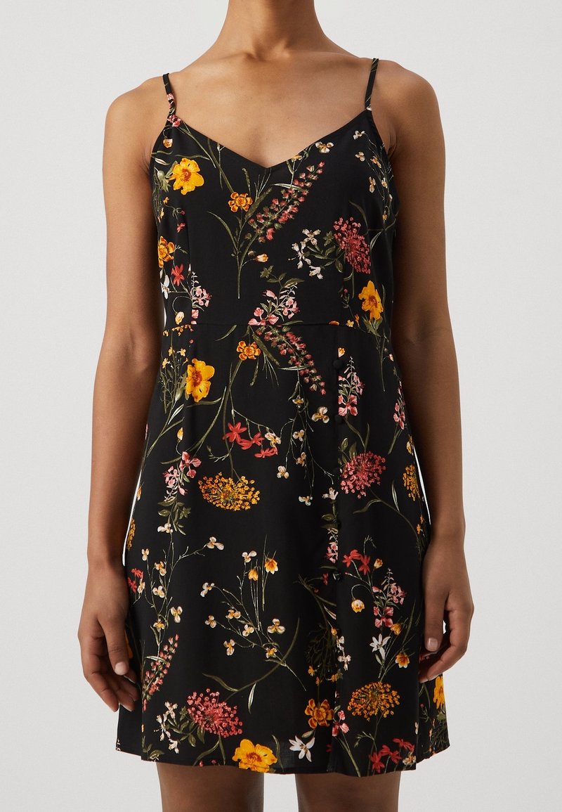 Femme portant une robe noire sans manches à imprimé floral multicolore et fines bretelles sur un fond clair uni.