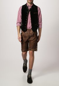 Schwarze Samtwesten über einem rot-weiß karierten Hemd, kombiniert mit braunen Ledershorts mit dekorativen Nähten und silbernen Knöpfen. Graue Socken und dunkle Schuhe.