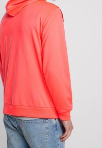 Sweat à capuche rose néon en tissu léger et lisse. Comprend une capuche, des poignets côtelés et un ourlet. Porté avec un jean déchiré bleu clair.