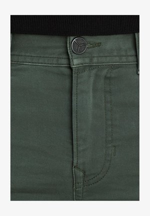 Vihreät farkkushortsit, joissa on vetoketjuluukku ja nappikiinnitys, etutaskut ja hillityt ommeltu yksityiskohdat. Materiaali on sileää ja leikkaus on muotoiltu.