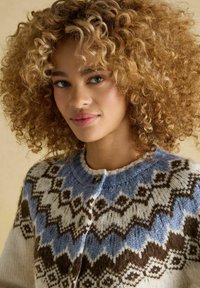 Kobieta z kręconymi blond włosami, ubrana w niebieski, biały i brązowy sweter dziergany ze wzorem i guzikami, spogląda nieco na bok.