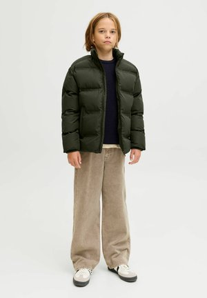 Chaqueta acolchada verde oscuro con cuello alto, que presenta un diseño acolchado. Llevada sobre un suéter azul marino y pantalones anchos de pana beige.