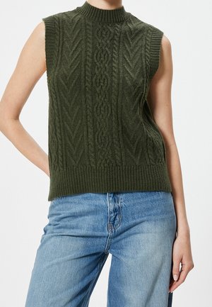 Gilet en maille torsadée vert foncé sans manches porté avec un jean en denim bleu clair sur une personne debout devant un fond uni.