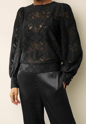 Personne portant un chemisier en dentelle noire à manches longues bouffantes et un pantalon en satin noir, main dans la poche.