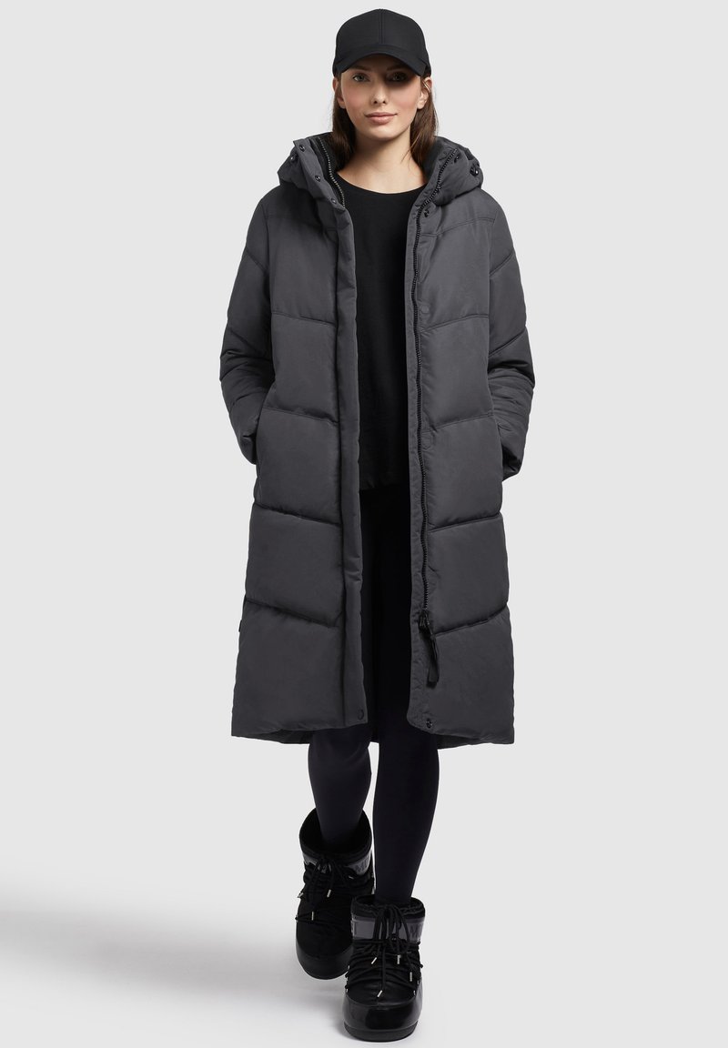 Cappotto lungo imbottito grigio scuro con cappuccio, dotato di chiusura con zip, tasche laterali e una texture liscia, abbinato a calzature nere.