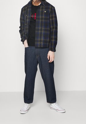 Skotsk flanneljakke i navy og gul, sort t-shirt med rødt logo, løstsiddende denimjeans og hvide sneakers.