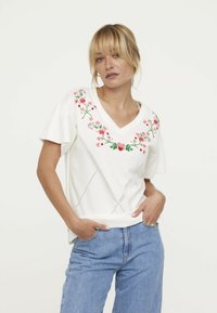 Haut à manches courtes blanc avec un col en V, mettant en avant des broderies florales colorées. Tissu texturé avec des détails de coutures diagonales et une coupe décontractée.