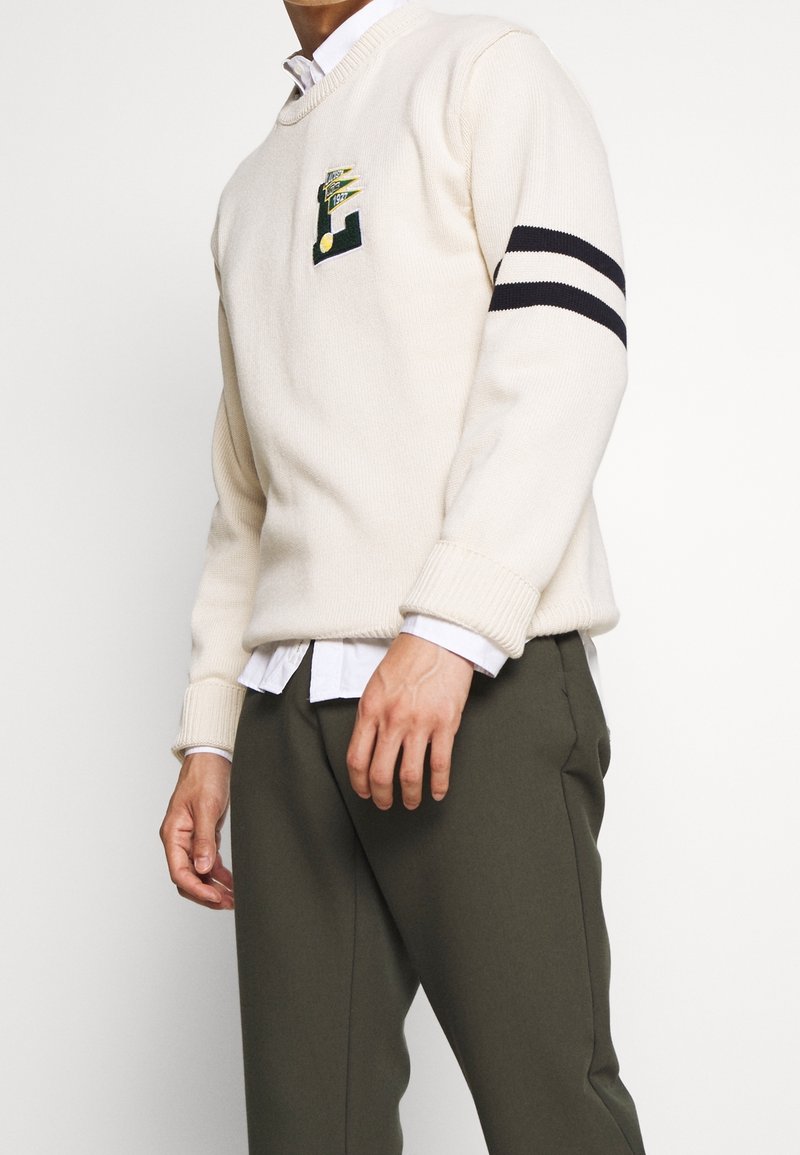 Krämfärgad tröja med rund halsringning och grön logopatch; har svarta ränder på ärmarna. Bärs över en vit skjorta med krage.