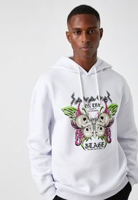 Sudadera blanca con un diseño gráfico colorido que incluye calaveras, alas de mariposa y texto. Tejido suave, cordones y puños acanalados.