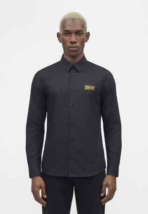 Mężczyzna z wybielonymi włosami, ubrany w dopasowaną czarną koszulę zapinaną na guziki z małym żółto-białym prostokątnym logo na lewej piersi.