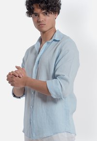 Camicia in lino azzurro chiaro con abbottonatura frontale, maniche lunghe arrotolate e vestibilità rilassata. Tessuto liscio con un design informale.