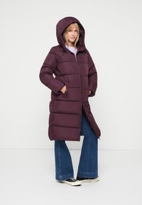 Dlhý, vypchávkovaný kabát v tmavofialovej farbe s kapucňou, s horizontálnym prešívaním, vreckami a zipsom. Model má na sebe rozšírené džínsy a tenisky.
