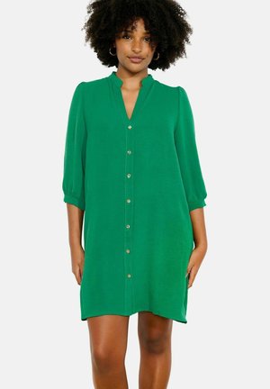 Ženska, oblečena v zelena obleka s kratkimi gumbi do kolen, z 3/4 rokavi in majhnim ovratnikom, stoji pred belim ozadjem.