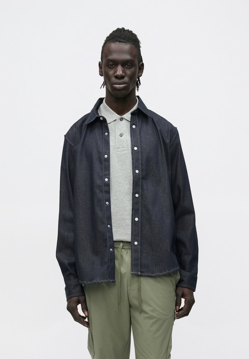 Granatowa koszula dżinsowa z zatrzaskami, casualowy kołnierzyk, noszona na szarym polo. Model prezentuje ją w połączeniu z jasnozielonymi spodniami. Gładka tekstura.