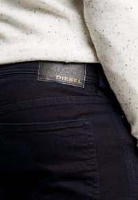Jeans de mezclilla azul oscuro con una etiqueta de cuero que presenta "DIESEL" en letras doradas en relieve en la parte trasera. Detalles de costura visibles.