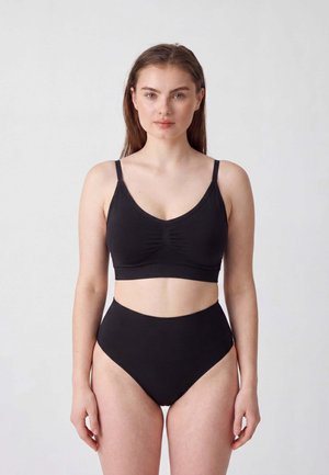Frau, die nach vorne blickend steht, trägt schwarze hoch taillierte Unterwäsche und ein schwarzes kurzes Tanktop vor einem schlichten hellen Hintergrund.