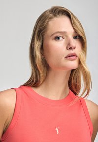 Koralowa bluzka bez rękawów z okrągłym dekoltem, wykonana z gładkiego materiału; posiada małe haftowane logo na piersi.