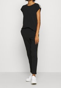 Czarny strój sportowy składający się z lekkiej bluzki z krótkim rękawem i dopasowanych spodni z kremowymi paskami po bokach. W połączeniu z białymi trampkami. Gładka tekstura.