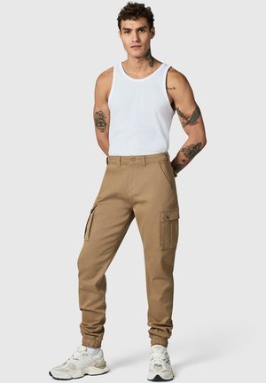Mężczyzna z tatuażami, ubrany w biały tank top, beżowe spodnie cargo jogger i białe trampki, stoi z rękami za plecami.